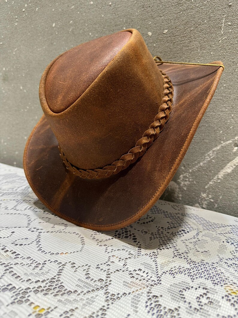 May include: A brown leather cowboy hat with a braided band around the crown. The hat has a wide brim and a slightly curved crown. The leather has a distressed look, giving it a vintage appearance.