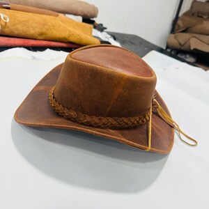 May include: A brown leather cowboy hat with a braided band around the crown and a wide brim. The hat has a distressed finish, giving it a vintage look. The hat is a classic western style.