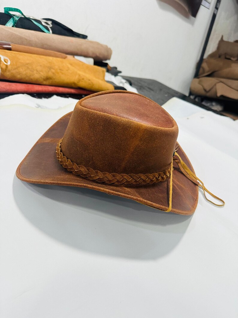 May include: A brown leather cowboy hat with a braided band around the crown and a wide brim. The hat has a distressed finish, giving it a vintage look. The hat is a classic western style.
