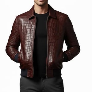 Peut inclure: Veste de bombardier en cuir marron avec une texture peau de crocodile. La veste a un col, une fermeture éclair et des poignets et un ourlet côtelés bleu foncé. La personne porte une chemise noire et un pantalon gris.