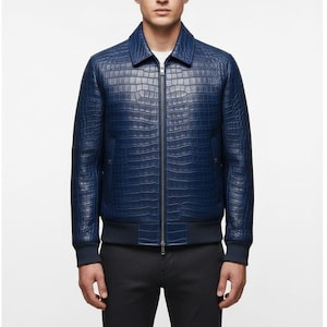 Może przedstawiać: Granatowa skórzana kurtka bomberka o fakturze skóry krokodyla. Kurtka ma kołnierz, zapięcie na zamek błyskawiczny oraz prążkowane mankiety i dół. Kurtka jest noszona na białej koszuli.