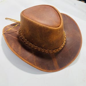 May include: A brown leather cowboy hat with a wide brim and a braided band around the crown. The hat has a distressed finish, giving it a vintage look. The hat is made of leather and has a classic cowboy hat shape.