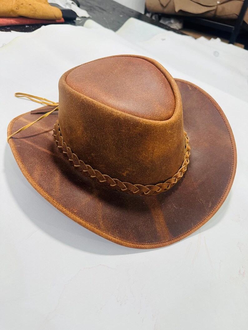May include: A brown leather cowboy hat with a wide brim and a braided band around the crown. The hat has a distressed finish, giving it a vintage look. The hat is made of leather and has a classic cowboy hat shape.