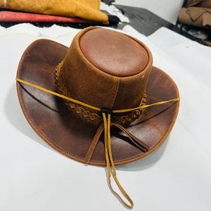May include: A brown leather cowboy hat with a braided band and a yellow leather chin strap. The hat has a rounded crown and a wide brim. The hat is made of distressed leather.