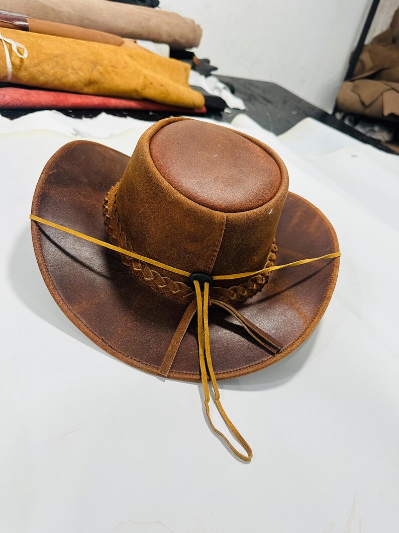 May include: A brown leather cowboy hat with a braided band and a yellow leather chin strap. The hat has a rounded crown and a wide brim. The hat is made of distressed leather.