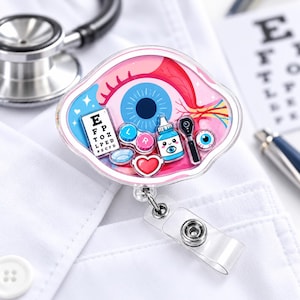 May include: A badge reel featuring a clear, eye-shaped acrylic casing filled with miniature charms including an eye chart with letters E, F, P, T, O, Z, L, P, E, D, P, E, C, F, D, contact lens cases, a blue eye drop bottle, an ophthalmoscope, and a red heart.