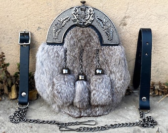 Puede incluir: Un sporran de piel gris y blanca con un adorno de metal plateado y negro con un diseño de cardo. El sporran tiene una correa de cuero negra y una cadena plateada.
