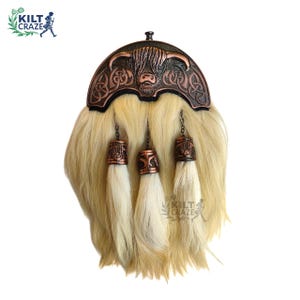 Puede incluir: Un sporran de kilt escocés tradicional con una parte superior de metal color cobre que presenta un diseño de vaca de las Tierras Altas y nudos celtas. El sporran tiene un cuerpo de piel de color claro y tres borlas decorativas.