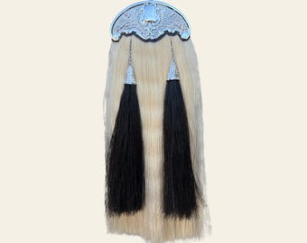 May include: A traditional Scottish sporran with a silver cantle featuring an ornate design. The sporran has a cream-colored fur body and two long, black tassels. The design is symmetrical and the tassels hang down from the bottom.