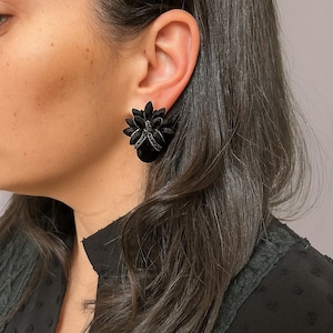 Könnte beinhalten: Schwarze Ohrstecker in Blumenform mit einer Ansammlung kleiner, dunkler Edelsteine. Die Ohrringe werden am Ohr einer Person getragen, die ein schwarzes Oberteil trägt. Die Ohrringe haben ein dunkles, elegantes Aussehen.