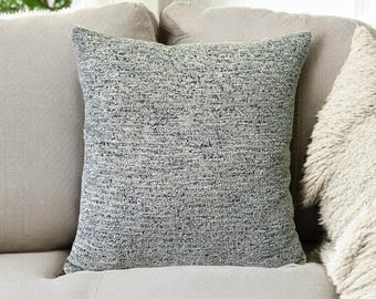 May include: A square, textured throw pillow in shades of gray and blue sits on a light gray sofa. The pillow has a woven appearance, adding a tactile element to the decor. The background includes a glimpse of a window and a fluffy, light-colored throw.