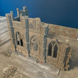 May include: A grey miniature model of a ruined gothic building. The model features arched windows, detailed stonework, and a partially collapsed structure, suggesting a war-torn or aged aesthetic. The model is set on a grey surface.