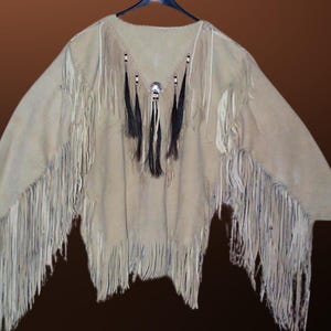 May include: A beige suede garment with fringe detailing and black feathers. The garment has a V-neck and is decorated with beads and a metal button.