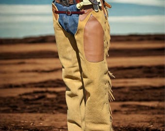 Può includere: Una persona indossa chaps in camoscio beige con toppe in pelle marrone e frange. I chaps sono indossati sopra jeans blu e una cintura marrone con fibbia argentata. La persona indossa anche stivali da cowboy marroni.