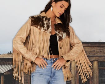 Könnte beinhalten: Beigefarbene Wildlederjacke mit langen Fransen und Kuhfell-Akzenten an Kragen und Schultern. Die Jacke hat eine Knopfleiste und lange Ärmel. Die Person trägt eine blaue Jeans und ein schwarzes Oberteil.