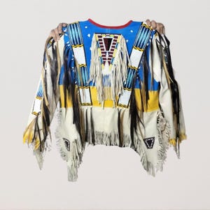 May include: A Native American-style shirt with a blue yoke and red neckline. The shirt features white, yellow, and blue geometric patterns, fringe, and black feather accents. The garment is held up by two hands.