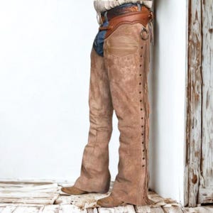 May include: Brown suede chaps with fringe and stud detailing. The chaps are worn with a brown leather belt and a denim and leather accent. The chaps have a classic western style.