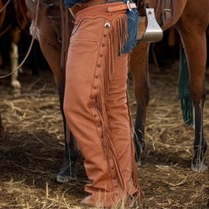May include: Brown leather chaps with fringe detailing and button closures. The chaps are worn over blue jeans and are secured with a brown leather belt. The image shows the chaps from the waist down, with a horse in the background.
