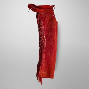 May include: Red suede chaps with fringe and white stitching. The chaps have a buckle closure at the waist.