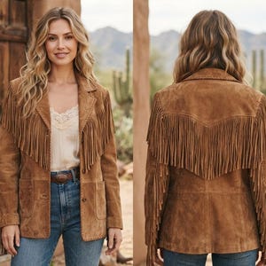 May include: A tan suede jacket featuring long fringe detailing across the chest, back, and sleeves. The jacket includes a notched collar, two front patch pockets, and button closures. The suede material has visible stitching along the edges.