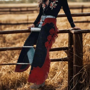 May include: Black and red fringed chaps with floral accents. The chaps are worn over blue jeans. The image also shows a black long-sleeve shirt with floral embroidery and brown cowboy boots. The background is a rustic fence and dry grass.