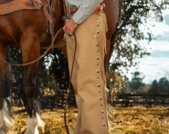 Può includere: Chaps in pelle color cuoio con frange e bottoni argentati. Gli chaps sono indossati sopra i pantaloni e fanno parte di un completo da equitazione western. Un cavallo e una sella sono visibili sullo sfondo, suggerendo un tema da ranch o rodeo.