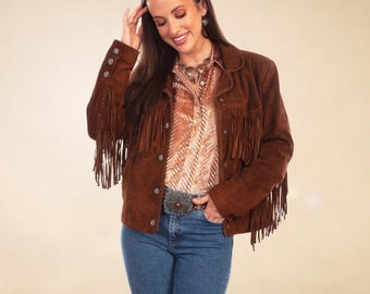 May include: A brown suede jacket with fringe detailing on the sleeves and chest. The jacket has button closures and is paired with a patterned button-down shirt and blue jeans. A decorative belt completes the look.