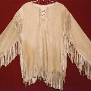 Peut inclure: Chemise en daim beige avec des manches longues et des franges. La chemise présente un décolleté à lacets et des franges le long des manches et de l'ourlet. La chemise est présentée sur un cintre sur fond rouge foncé.