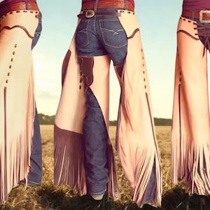 May include: Tan leather chaps with fringe detailing, worn over blue jeans. The chaps feature decorative stitching and a brown leather belt. The image shows the chaps from multiple angles, showcasing the design and texture.