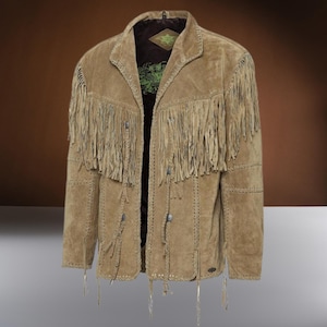 Puede incluir: Chaqueta de ante color canela con largos flecos en el pecho y las mangas. La chaqueta tiene cuello, costuras decorativas y detalles trenzados. Cuenta con botones plateados y un pequeño logotipo cerca del dobladillo. Debajo se ve una camisa oscura con un gráfico verde.