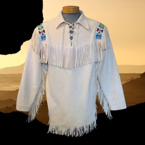 May include: Cream-colored suede shirt with long sleeves and fringe detailing. The shirt features colorful beaded shoulder accents and a laced neckline. The fringe is along the chest, sleeves, and hemline. The shirt is displayed on a mannequin.
