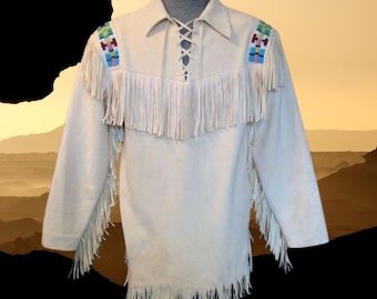 May include: Cream-colored suede shirt with long sleeves and fringe detailing. The shirt features colorful beaded shoulder accents and a laced neckline. The fringe is along the chest, sleeves, and hemline. The shirt is displayed on a mannequin.