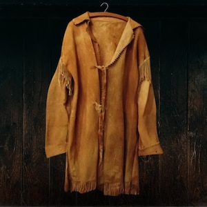 Könnte beinhalten: Eine braune Lederjacke mit Fransenbesatz. Die Jacke hängt an einem Holzbügel vor einem dunklen Holzhintergrund.