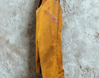 Può includere: Chaps in pelle giallo senape con cuciture decorative e frange sui lati. Gli chaps presentano una cintura e dettagli in pelle marrone, con un anello e fibbia in metallo. Esposti contro un muro grigio strutturato e pavimento in legno.