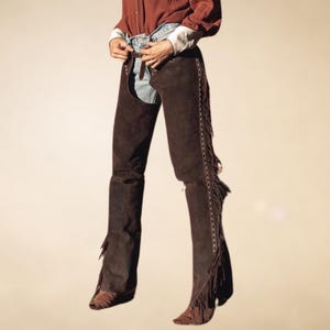 May include: Brown leather chaps with fringe detail and a white stitching design. The chaps are worn over blue jeans.