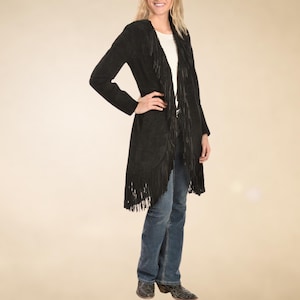 May include: A long black suede jacket with fringe detailing along the hem and lapels. The jacket is open-fronted and worn over a white top and blue jeans. The jacket has long sleeves and a relaxed fit.