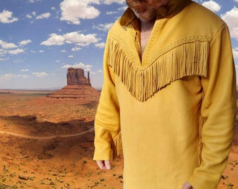 Puede incluir: Camisa de ante amarillo mostaza con mangas largas y diseño de cuello en V. La camisa presenta flecos decorativos a lo largo del escote y un cuello marrón en contraste. El fondo muestra un paisaje desértico con cielo azul.