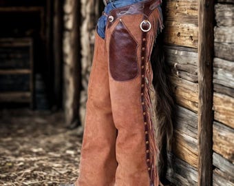 Può includere: Chaps in pelle marrone con frange e dettagli marrone scuro. Gli chaps hanno una grande toppa marrone scuro sulla coscia e sono decorati con piccole borchie lungo il lato. Gli chaps sono indossati con una cintura e jeans.