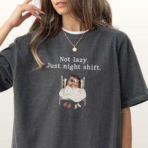 May include: Dark grey t-shirt with the text "Not lazy. Just night shift." and a cartoon hedgehog in bed. The shirt has a relaxed fit and short sleeves. The brand name "Color Pepper" is printed at the bottom left.