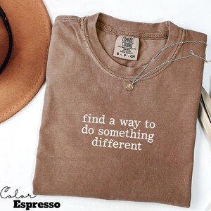 May include: Espresso-colored t-shirt with the text "find a way to do something different" in white. A silver necklace with a small pendant rests on the shirt. The shirt is folded on a white surface.