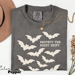 May include: A dark gray t-shirt with a bat graphic and the text "PROTECT THE NIGHT SHIFT". The shirt has a crew neck and short sleeves. A gold necklace with a star pendant is draped across the shirt.