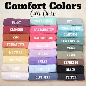 Pode incluir: Um gr&aacute;fico de cores intitulado "Comfort Colors" exibe uma variedade de amostras de tecido dobradas. As cores incluem Berry, Crimson, Yam, Terracota, Mostarda, Banana, Marfim, Branco, Island Reef, Crunchberry, Watermelon, Blossom, Orchid, Violeta, Chambray e Blue Jean.