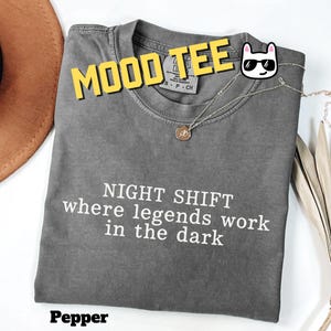 May include: A charcoal gray short-sleeve t-shirt with the white text "NIGHT SHIFT where legends work in the dark" printed on the front. The shirt features a crew neckline and a tag that reads "Comfort Colors" with a size S-CH label.