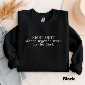 May include: Black crewneck sweatshirt with the text "NIGHT SHIFT where legends work in the dark" in white. The word "Black" is printed in the bottom right corner. The sweatshirt is displayed on a light background.