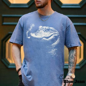 May include: Blue short-sleeve t-shirt with a white graphic of an alligator smoking a cigarette. The shirt has a relaxed fit and is made of a soft material. The text "Color Blue Jean" is visible on the bottom right.