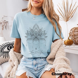 Pode incluir: Camiseta azul claro com um design detalhado de flor de l&oacute;tus preta e a frase "Love You". A camiseta &eacute; na cor chambray e tem mangas curtas. O tema geral &eacute; espiritual e bo&ecirc;mio.