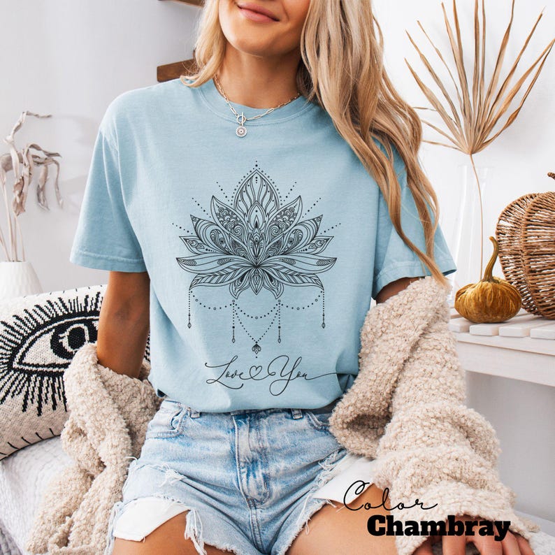 Pode incluir: Camiseta azul claro com um design detalhado de flor de l&oacute;tus preta e a frase "Love You". A camiseta &eacute; na cor chambray e tem mangas curtas. O tema geral &eacute; espiritual e bo&ecirc;mio.