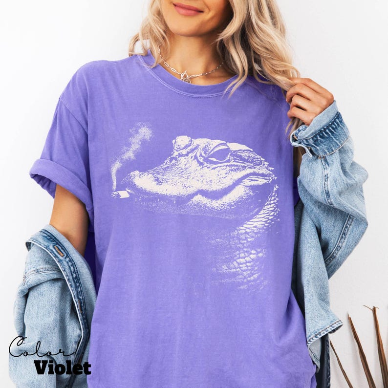 May include: A purple t-shirt featuring a white graphic of an alligator smoking a cigarette. The text "Color Violet" is printed on the bottom left of the shirt. The shirt has rolled-up sleeves and is worn with a denim jacket.