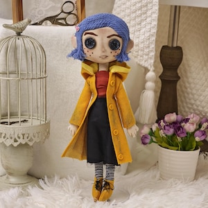 K&ouml;nnte beinhalten: Handgefertigte Puppe, inspiriert von der Figur Coraline. Die Puppe hat blaue Haare, Knopfaugen und tr&auml;gt einen gelben Mantel, ein rotes Oberteil und einen schwarzen Rock. Die Puppe steht auf einer wei&szlig;en Oberfl&auml;che.