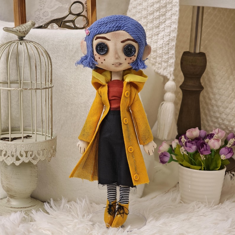 K&ouml;nnte beinhalten: Handgefertigte Puppe, inspiriert von der Figur Coraline. Die Puppe hat blaue Haare, Knopfaugen und tr&auml;gt einen gelben Mantel, ein rotes Oberteil und einen schwarzen Rock. Die Puppe steht auf einer wei&szlig;en Oberfl&auml;che.